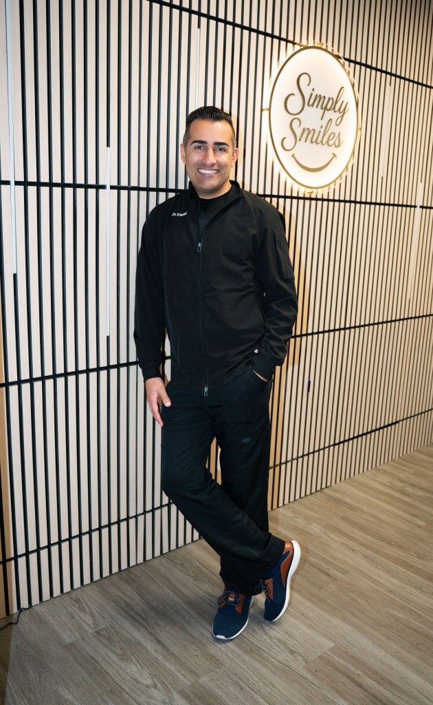 Dr. Amir Karimi leaning against his office wall smiling in black scrubs